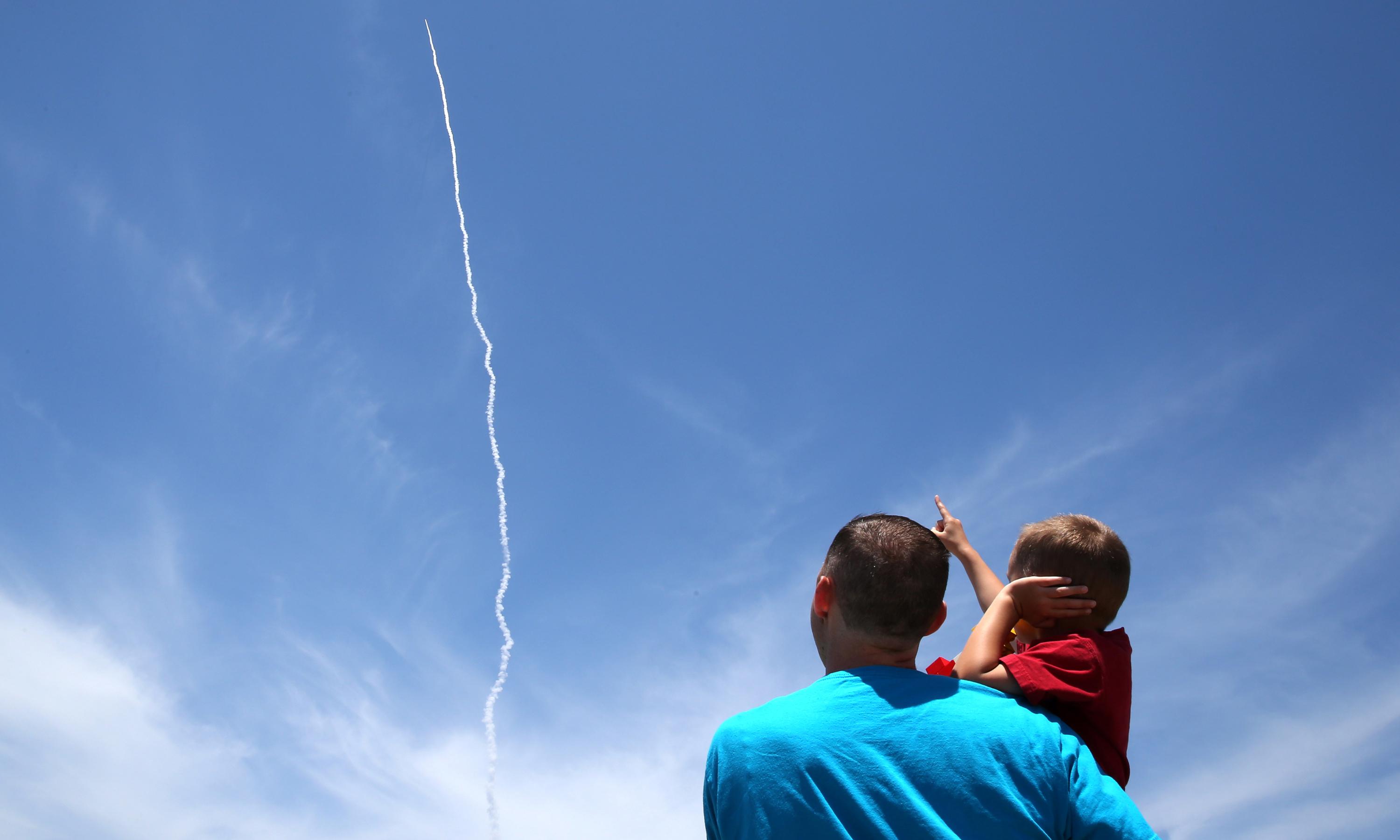A young boy held by his father points at the smoke trail of a missile in the sky.