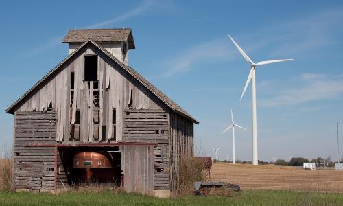 A horse trailer housed in a distressed barn situated in a field with wind turbines
