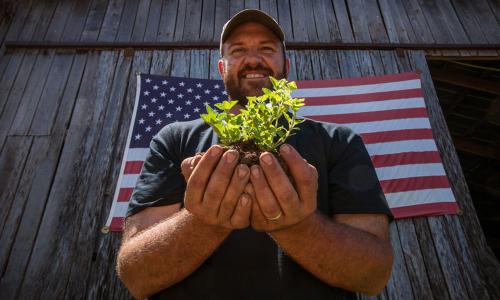 Farmer holding small plant in ball of soil in front of US flag