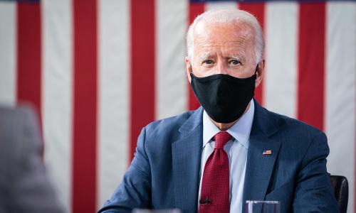Photo of presidential candidate Joe Biden sitting with a mask on and a flag-like red and white background