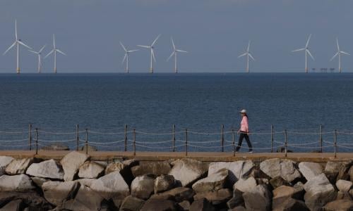 A person walks down a path with offshore wind turbines visible in the background.