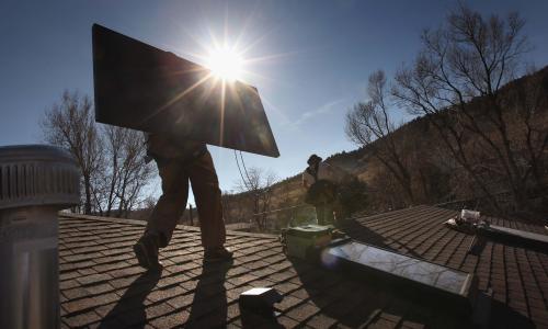 Two people install rooftop solar panels on a residential building.