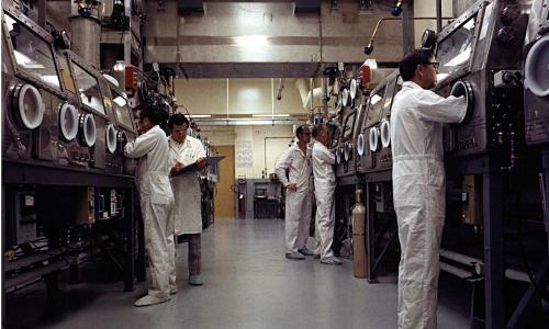 photo of several scientists in lab coveralls standing in front of two rows of glove boxes, with their hands inside the gloves so they can work with plutonium