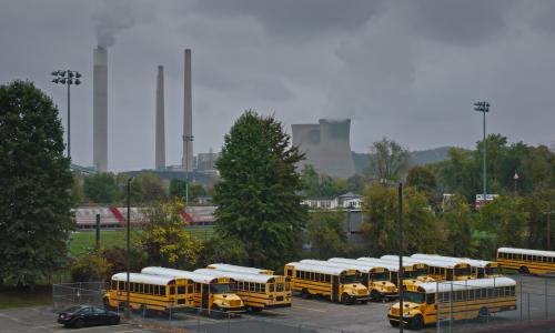 A line of school buses in front of smoke stacks.